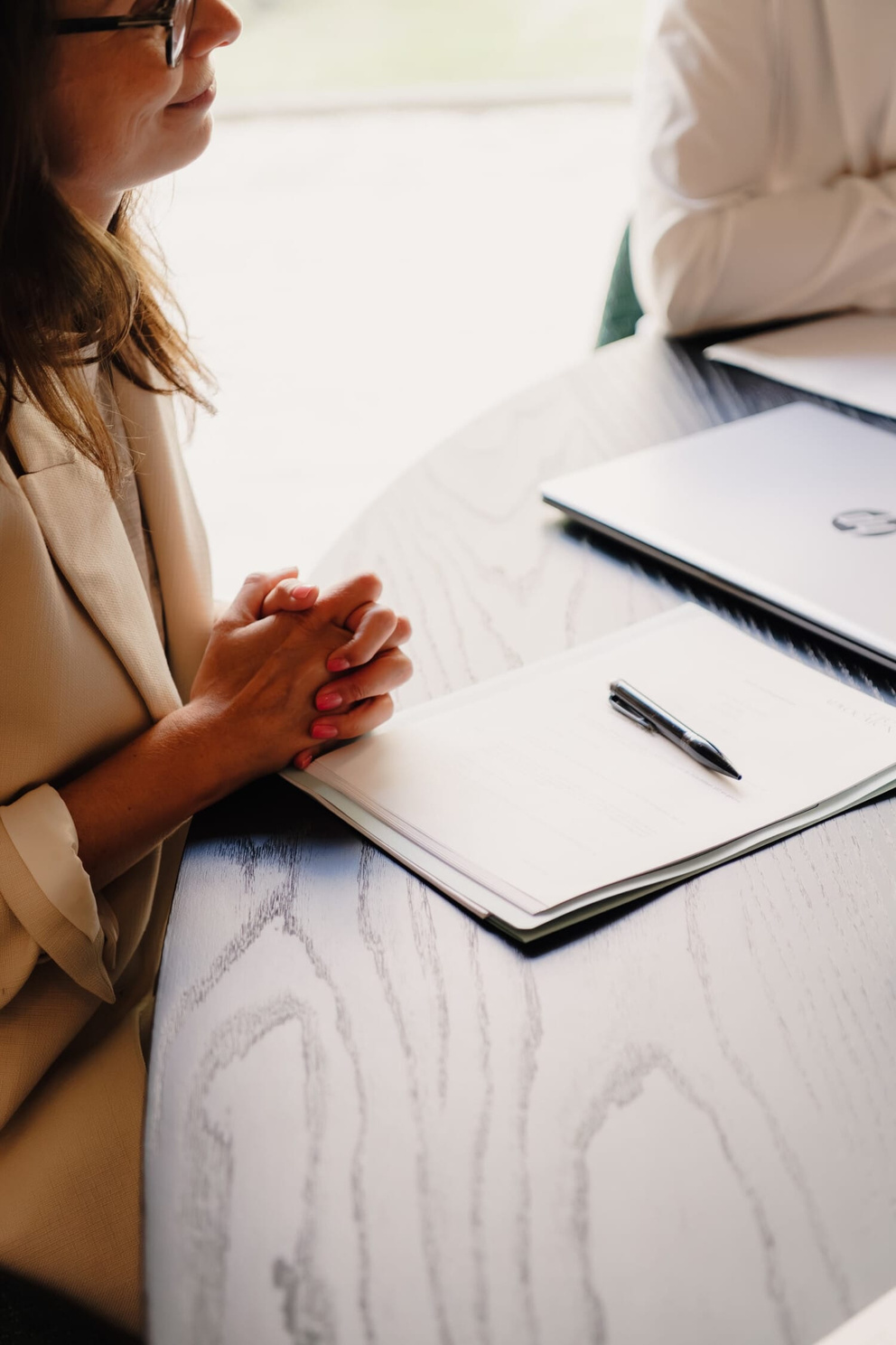 Een vrouw in een beige blazer met gevouwen handen aan een houten tafel, met een notitieboek en pen ervoor.