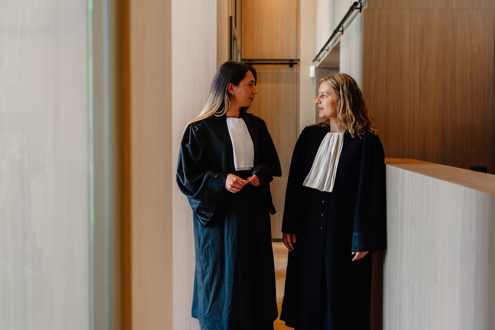 Twee vrouwelijke advocaten in toga's staan in een moderne rechtszaal en praten met elkaar.