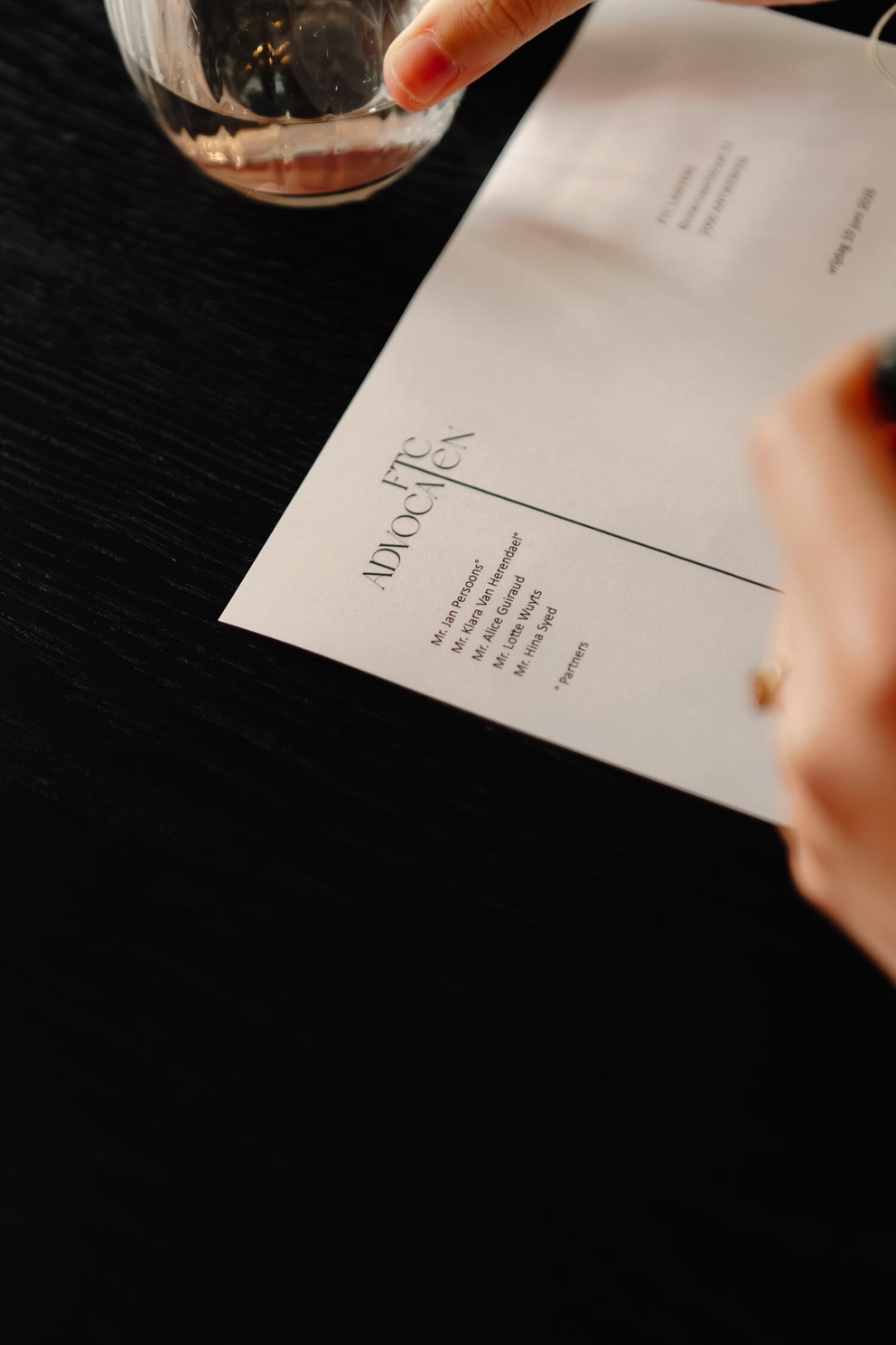 Een hand schrijft met een pen op een blanco brief met een elegante bovenkant en een glas water op de achtergrond.