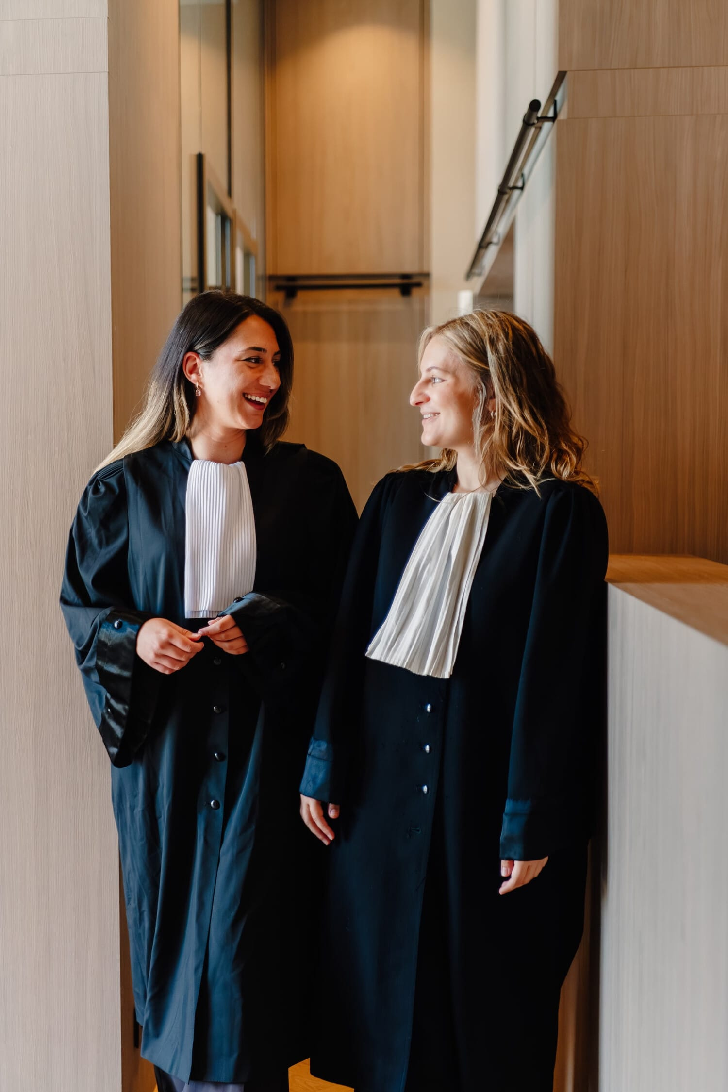 Twee vrouwelijke advocaten in toga's lachen naar elkaar in een moderne kantoortuin.