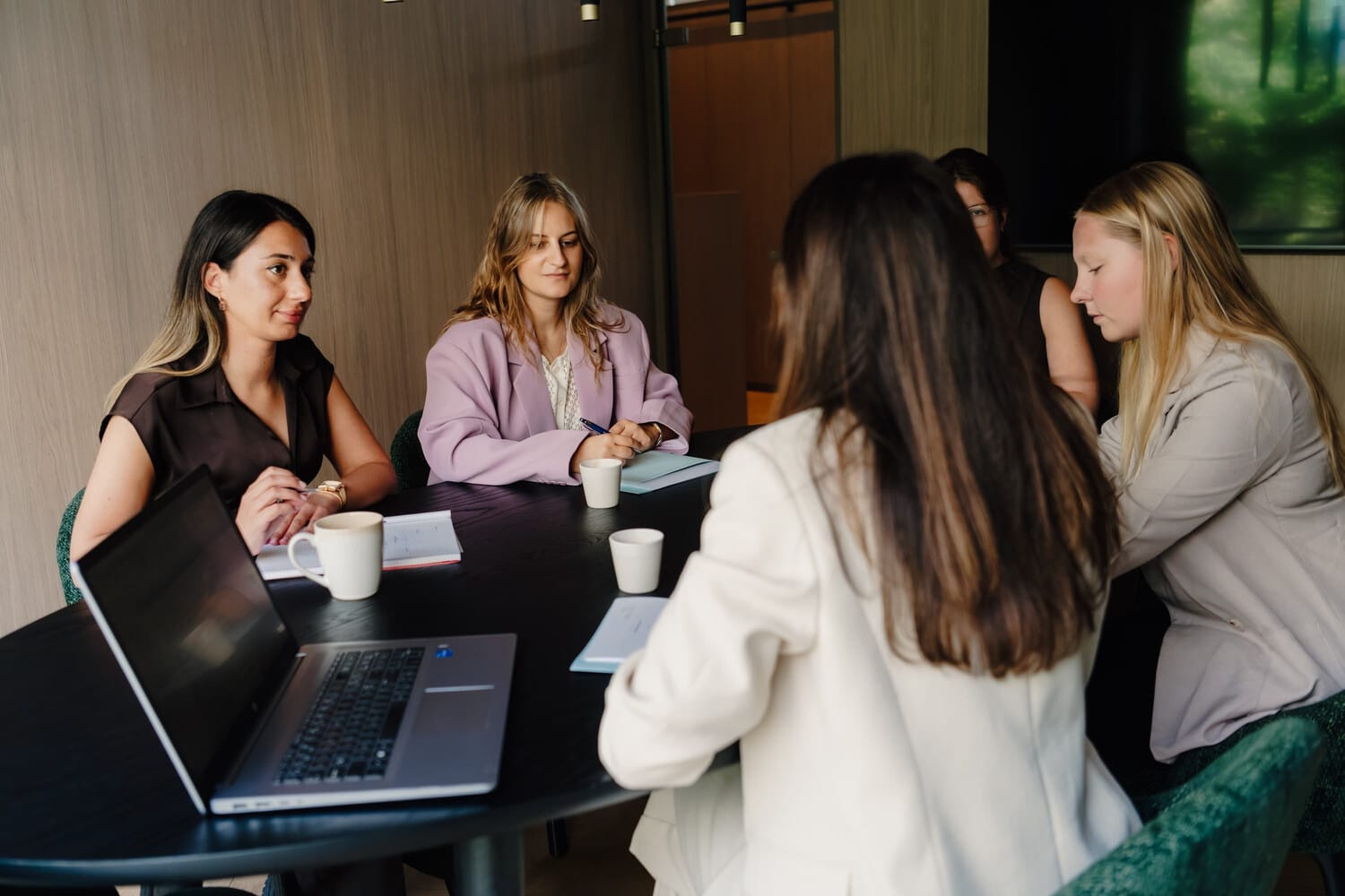 Vijf vrouwen in zakelijke kleding zitten rond een tafel met laptops en notitieblokken in een moderne vergaderruimte.