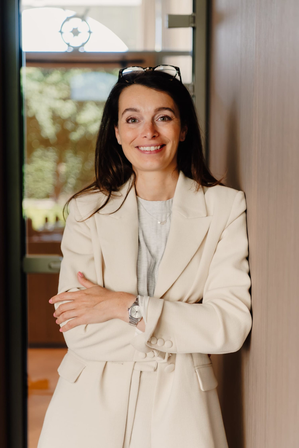 Vrouw in een lichte blazer met een vriendelijke glimlach, leunt ontspannen tegen een wand in een moderne omgeving.