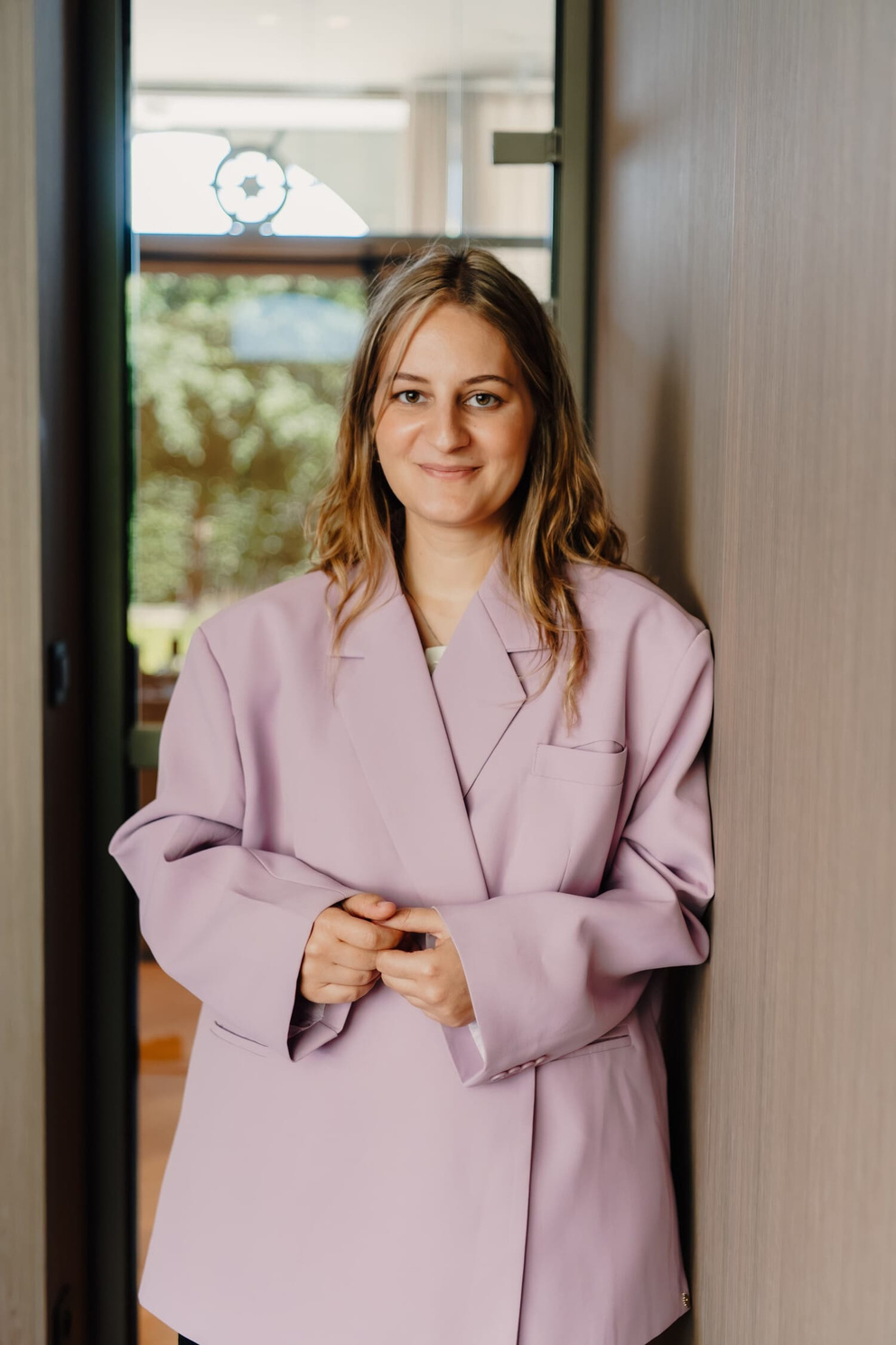 Jonge vrouw met halflang, golvend haar in een lila oversized jasje, staand bij een deuropening met een houten wand achter zich.